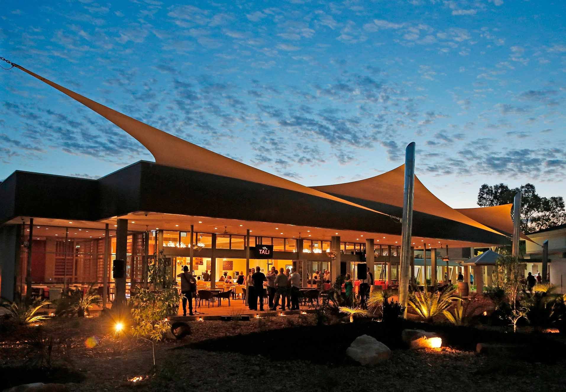 Modern building with large sail-like canopies, lit warmly at dusk. People gather inside and outside, surrounded by landscaped gardens.